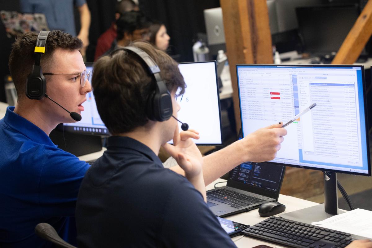 Seated at their terminals in mission control, Mason Starr points at Conner Awald's computer screen with his pen while Awald looks on.