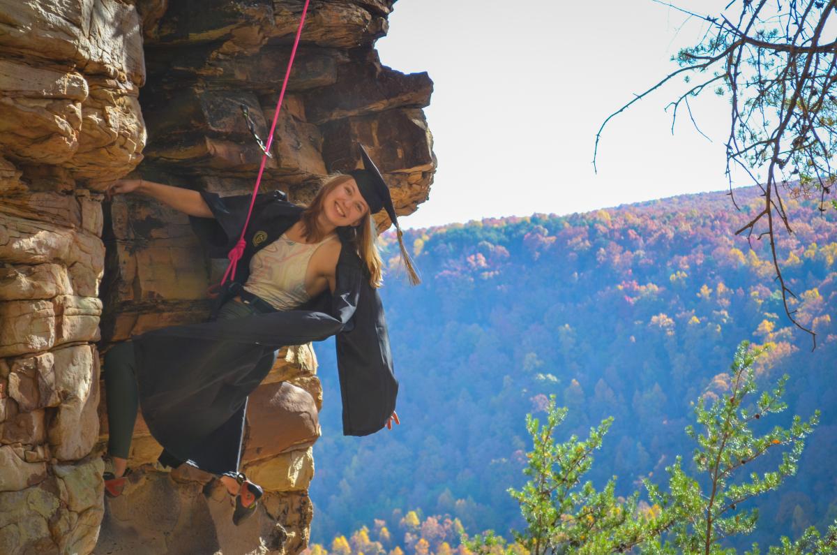 Mackenzie Sicard hangs from a cliff in rock climbing gear in her commencement regalia
