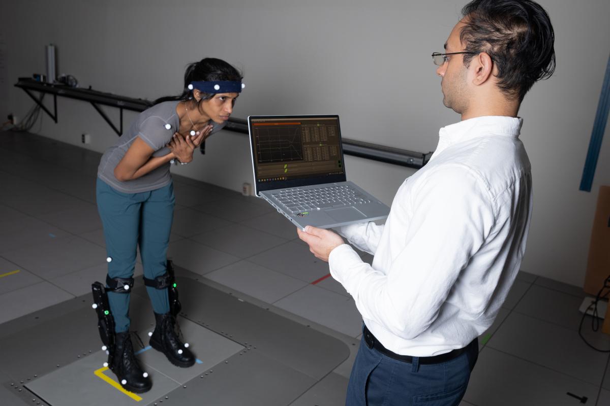 a woman wearing ankle exoskeleton boots stands on a moveable floor while a man monitors on a laptop