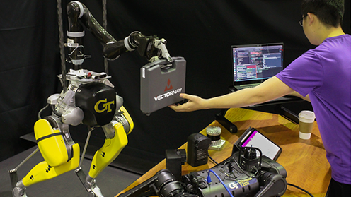 A two-legged robot with a manipulator arm holds a case while a researcher in purple shirt reaches for the case.