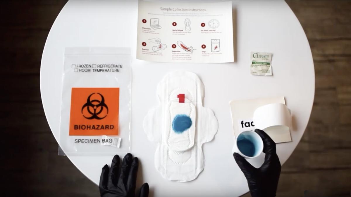 A round table with the FADpad kit: a menstrual pad with the FADpad add-on — with blue-colored liquid on the pad — a biohazard bag, instruction page, alcohol wipe, and a hand holding a small cup of blue sample liquid.
