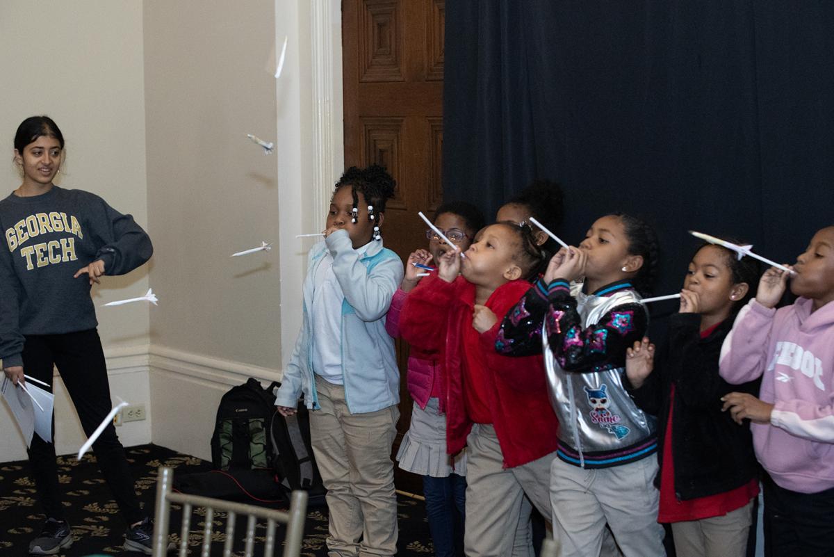 Elementary school students blowing through straws to "launch" the paper rockets they designed while a college student in Georgia Tech sweatshirt looks on. (Photo: Veronica Soroka)