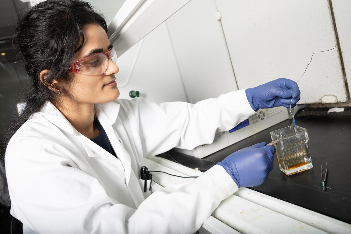 A researcher in lab coat, glasses, and gloves, positions electrodes above a small glass chamber. She's examining a small piece of stainless steel connected to one of the electrodes. (Photo: Candler Hobbs)
