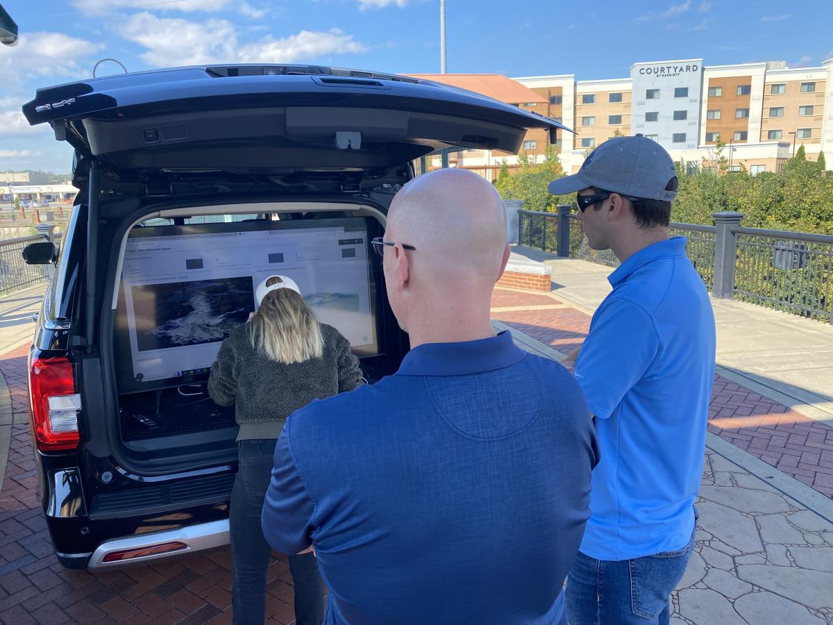 Three people look at a monitor in the back of an SUV