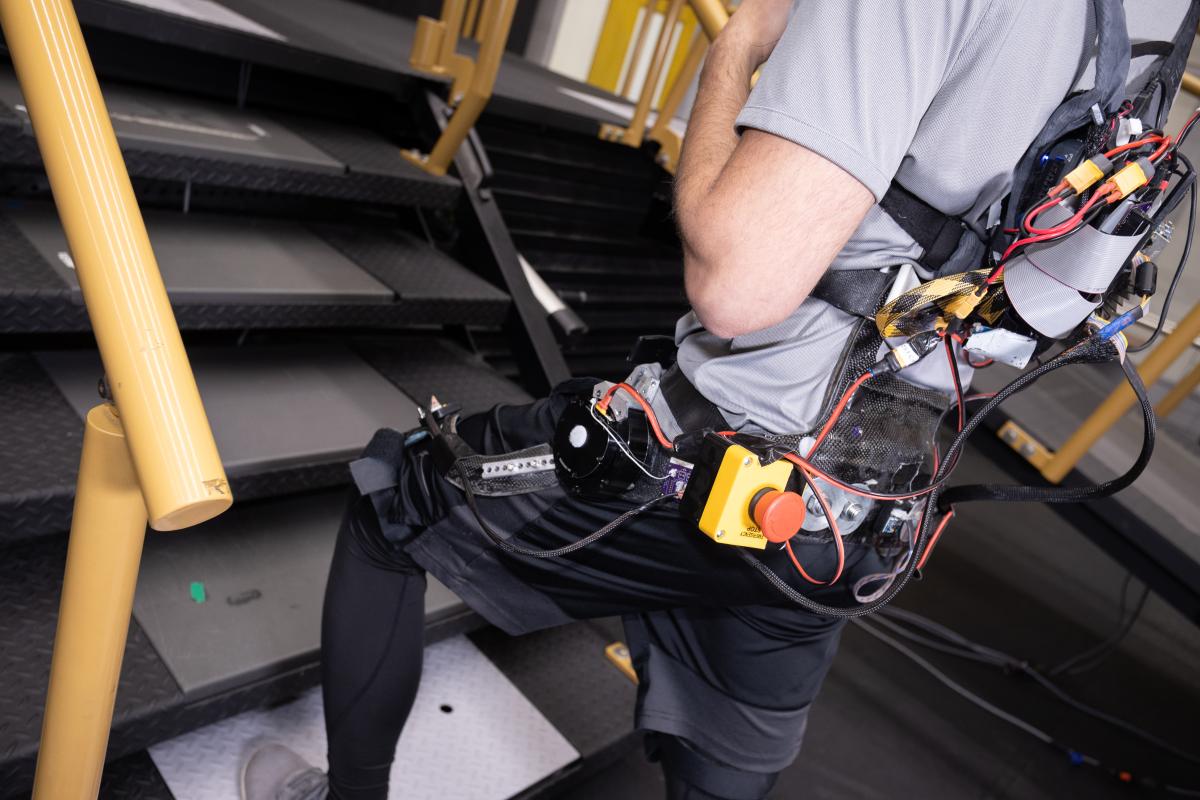 a person wearing a robotic assistive device on his back and legs climbs a set of stairs in a lab 