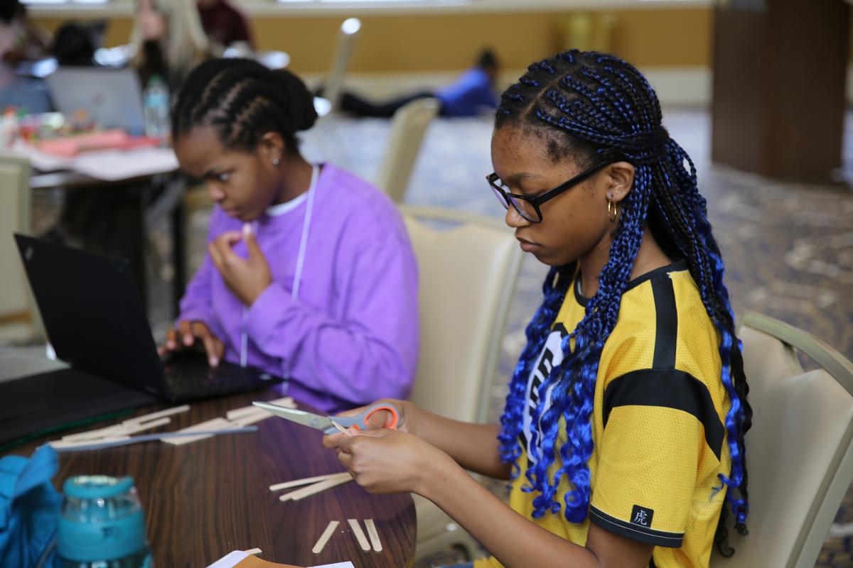 Two young students work on a project at a table. One is cutting materials with scissors and one is looking at a laptop. (Photo: Mikey Fuller)