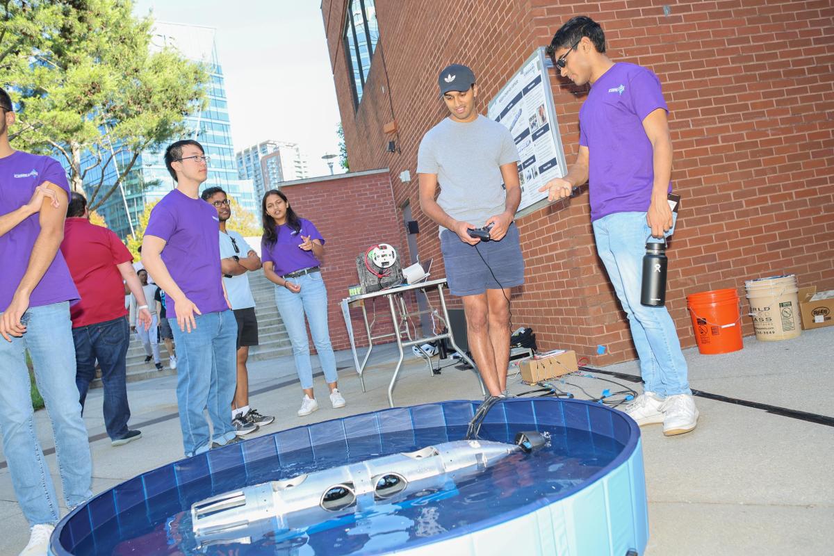 Students look at a long, cylindrical robot in a small kiddie pool of water at the Capstone Design Expo in spring 2024.