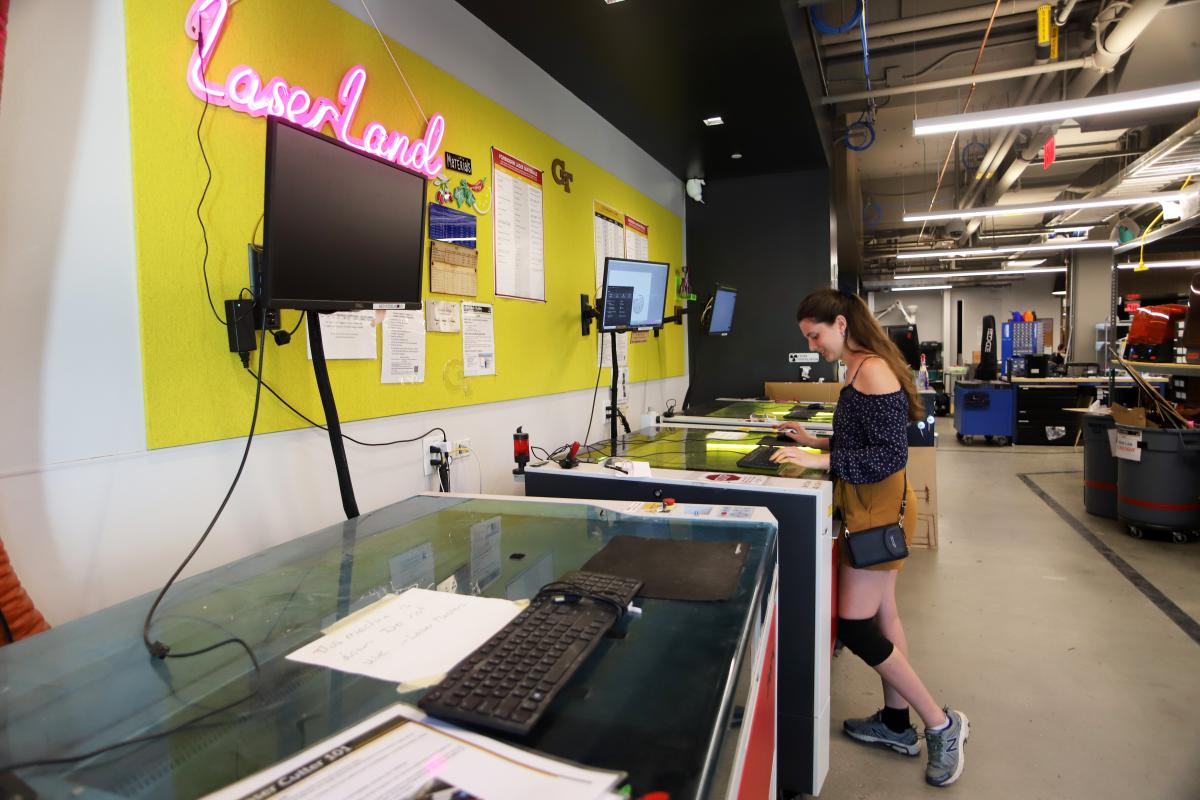 A student works at a laser cutter in the Flowers Invention Studio with a neon "Laser Land" sign on the wall.