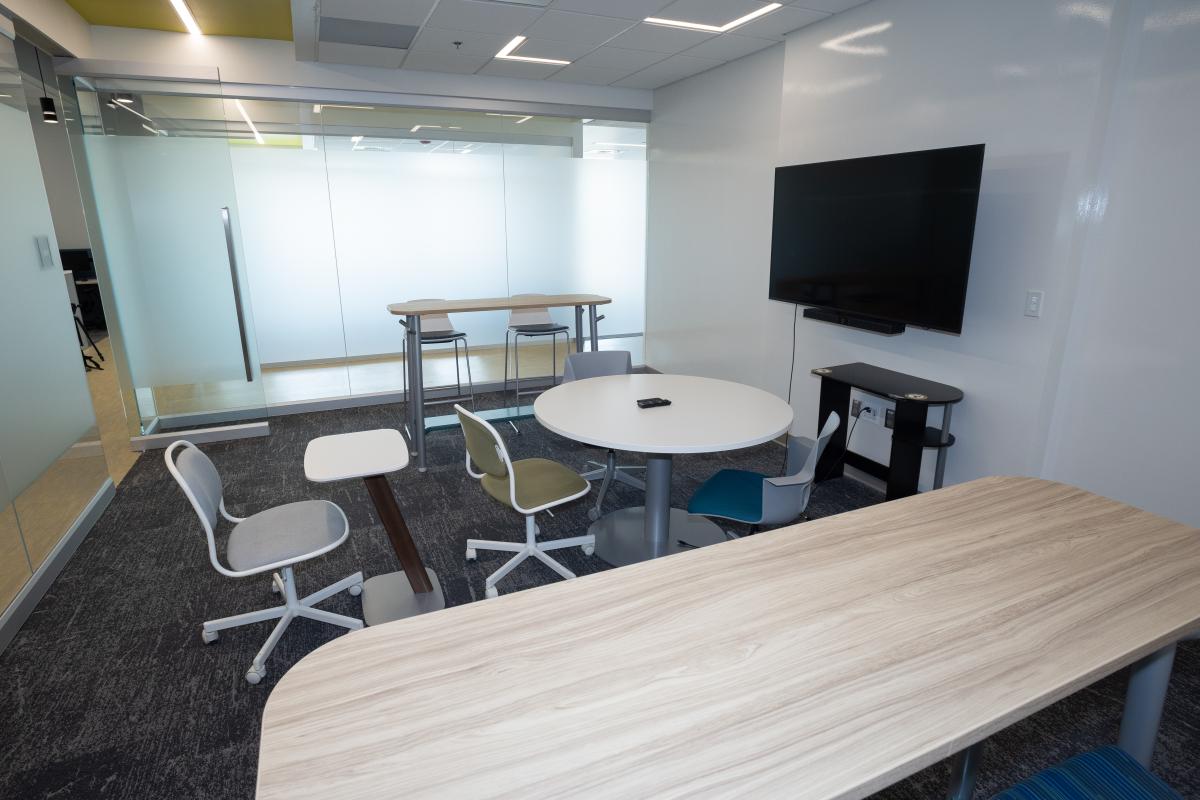 A renovated huddle room in the Tech Way Building with movable furniture and glass walls. A large display screen is mounted on the wall.