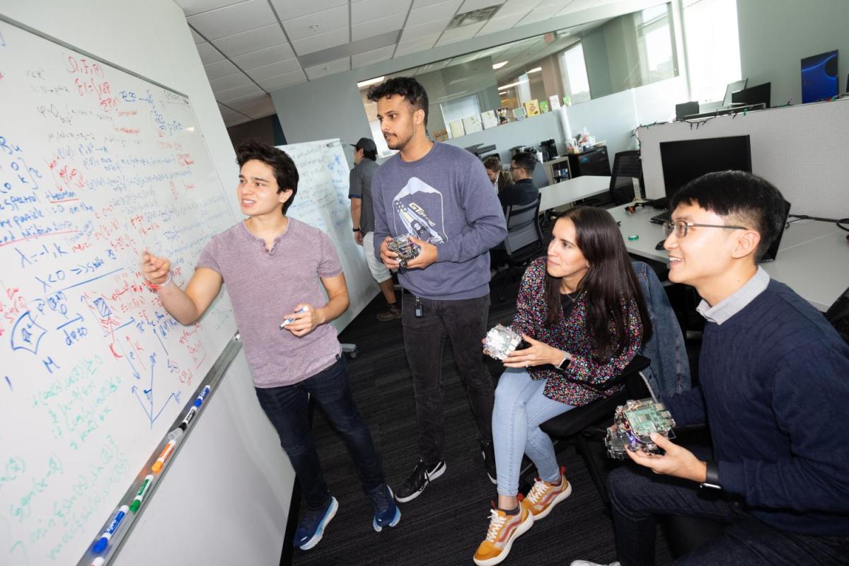 Four students holding robots look at equations on a whiteboard