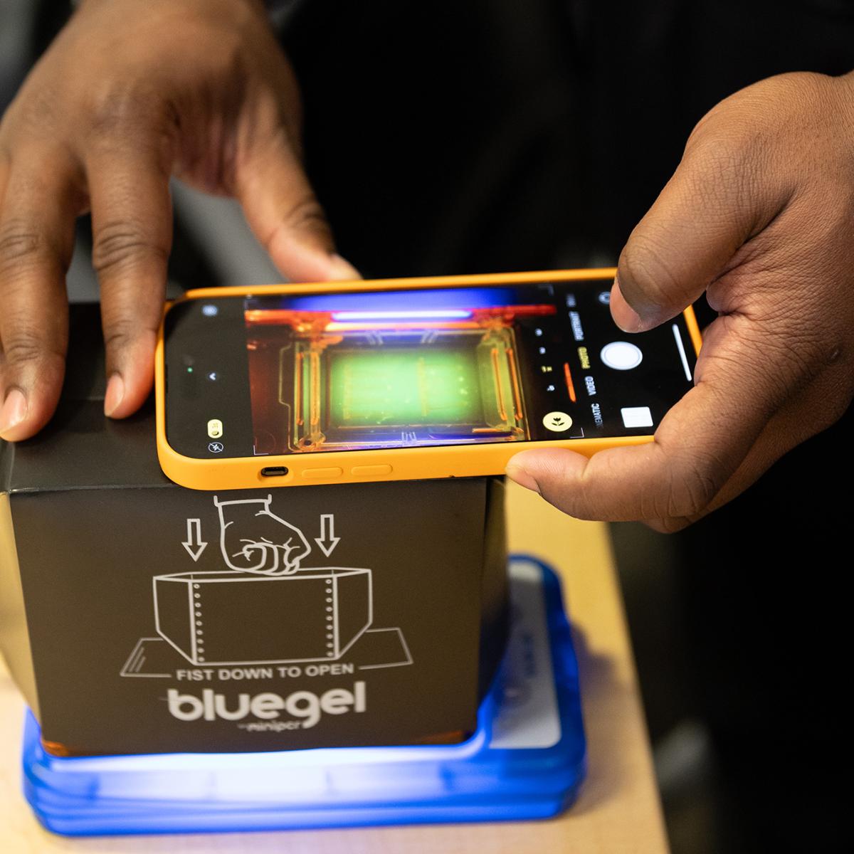 Two hands holding a smartphone over a box sitting on a small blue platform. On the screen, the camera app is showing a biological sample in the box. (Photo: Jihoon Kim)