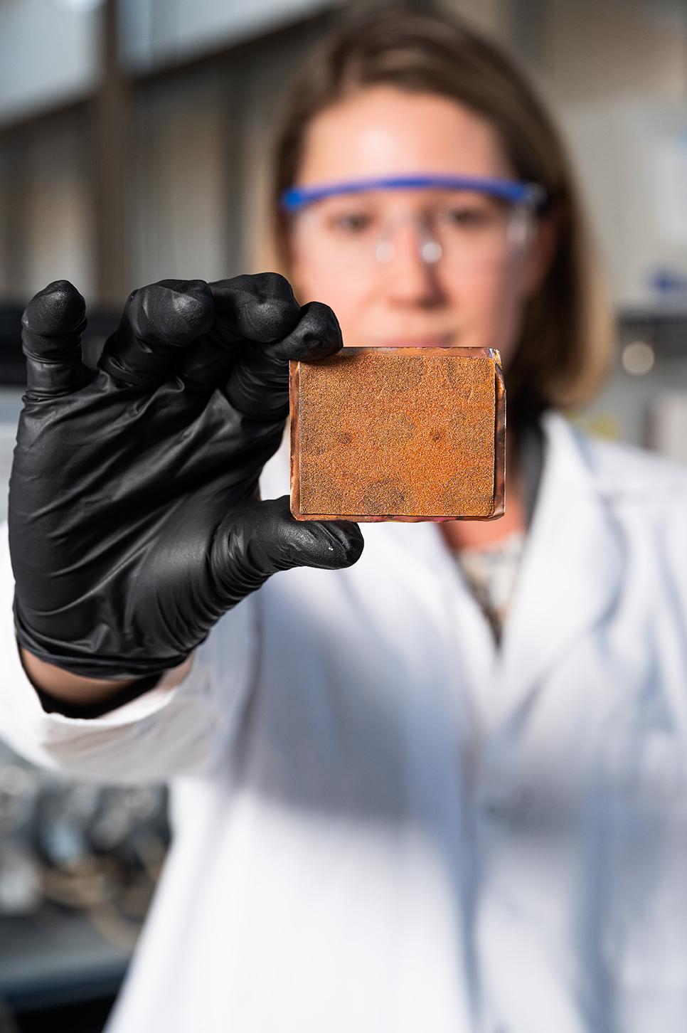 A female researcher in lab coat, safety glasses, and gloves holds an orange rectangular material out toward the camera. (Photo: Joya Chapman)