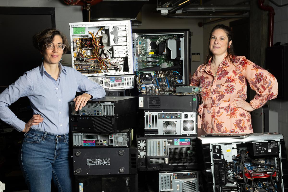 two women stand next to a pile of computers, circuitboards, and other electronic waste