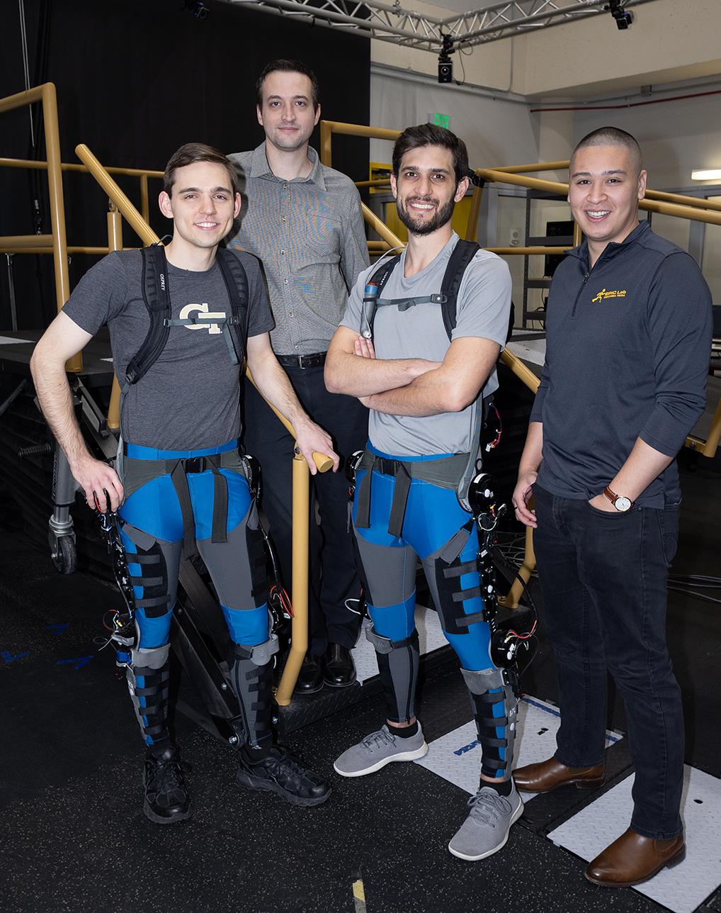 Four men standing in a lab. Two in the front are wearing robotic exoskeleton devices on their legs.