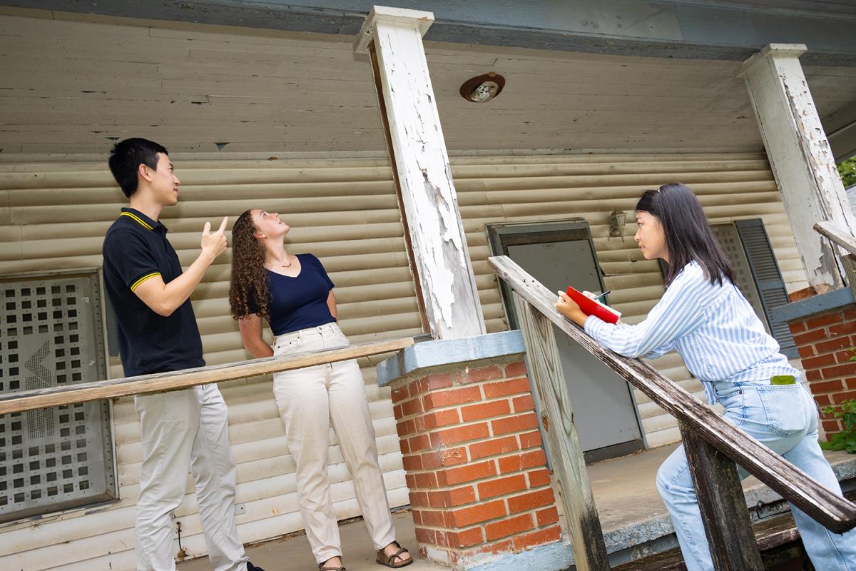 Three students look at a house's structure from the front porch