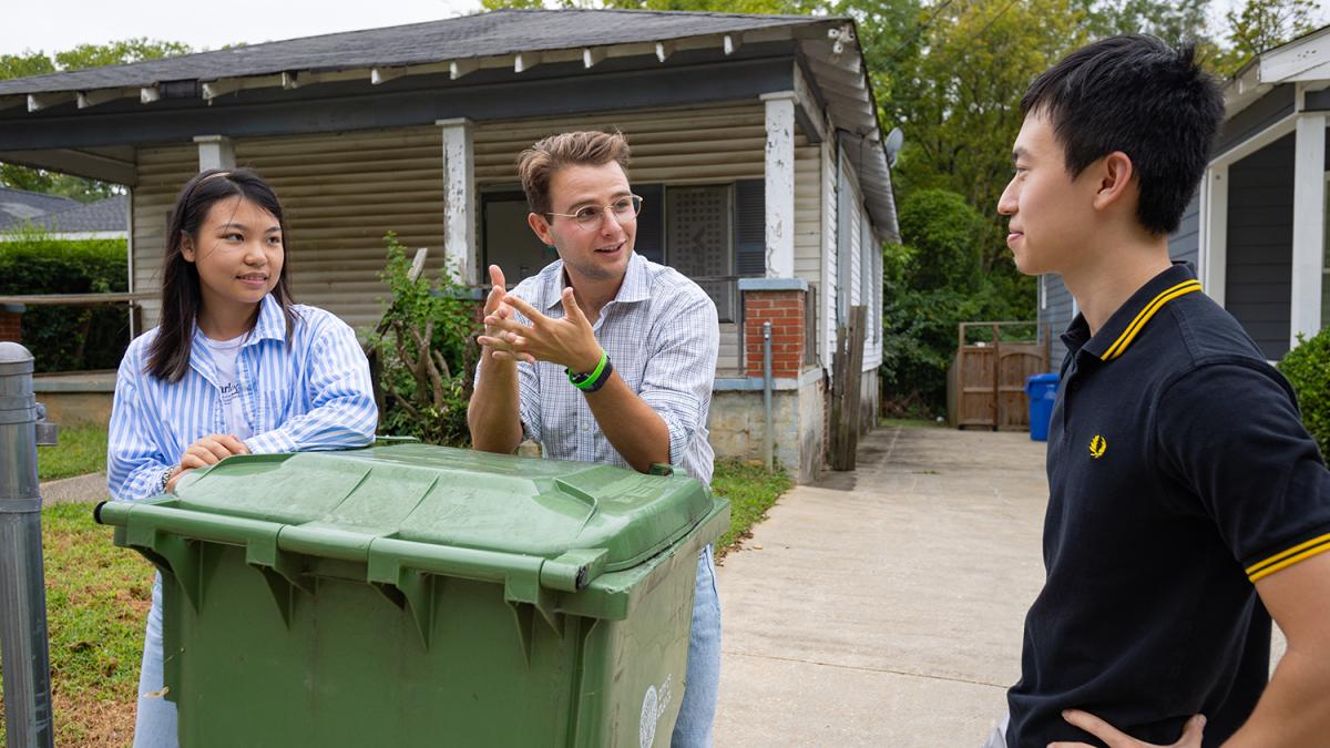 three students talk next to a trashbin in front of a house in Vine City