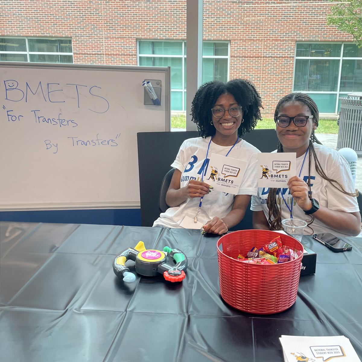 Two women sitting at a table and holding flyers. A whiteboard beside them reads BMETS "For transfer students by transfer students"