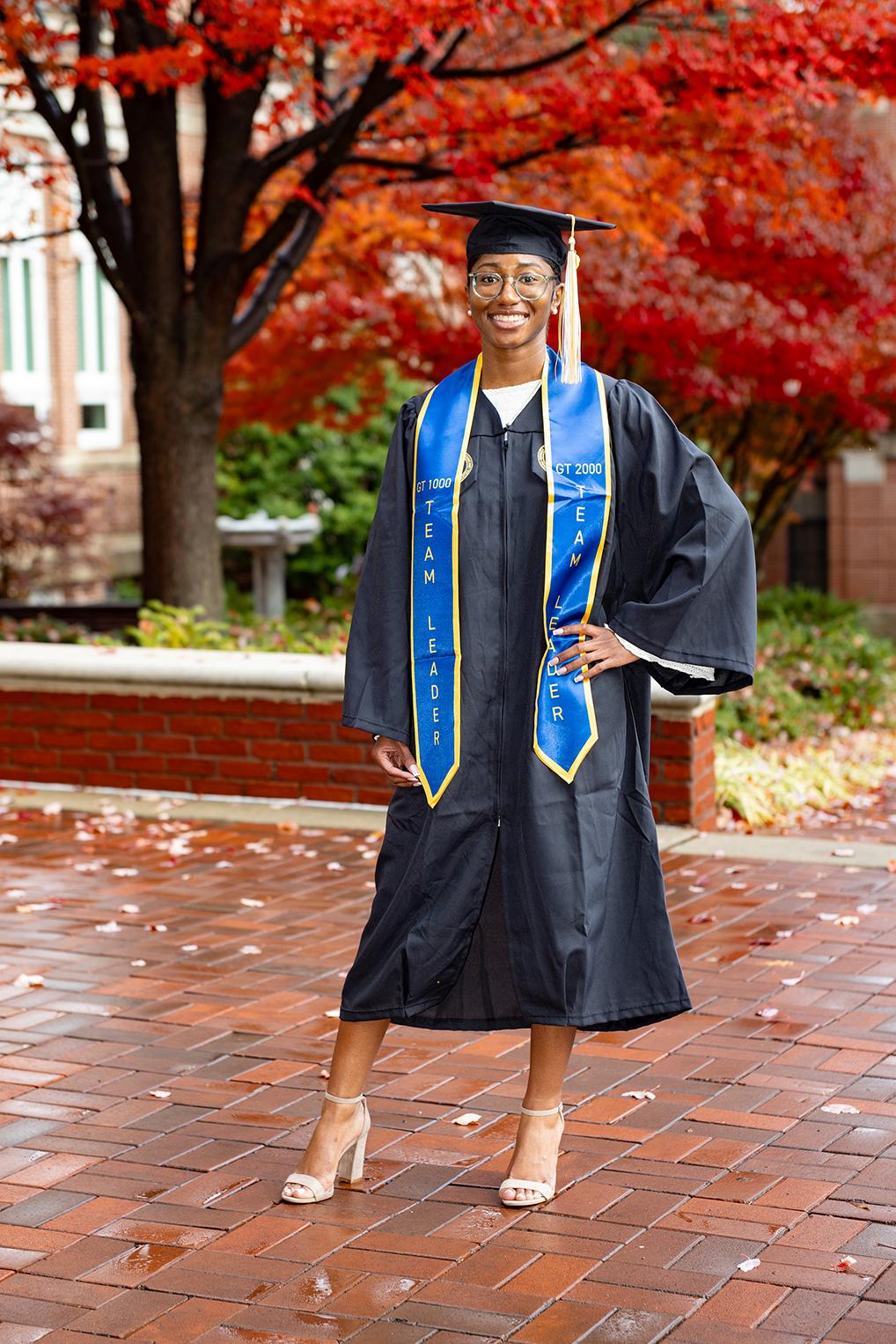 Afeni Laws stands in her cap and gown on a brick walkway with a deep orange and red tree in the background.