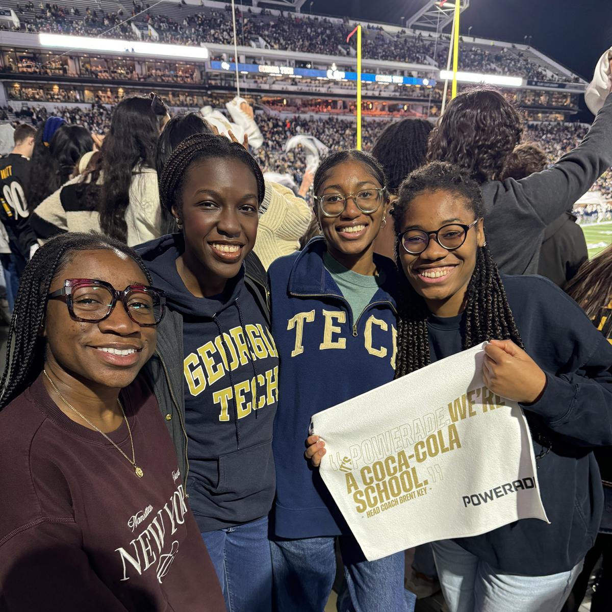 Four women smiling at the camera at a football game with the stadium and goal posts in the background.