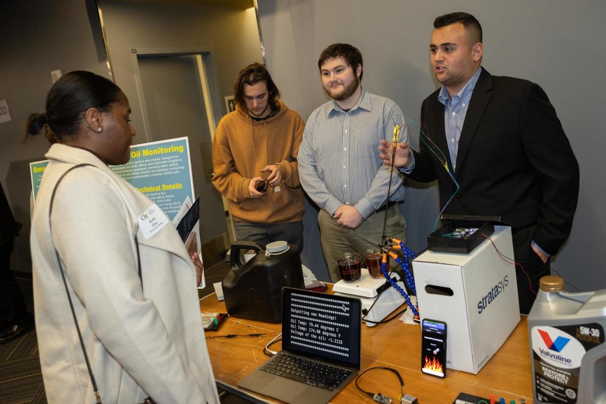 three male students talk to an attendee while standing behind computers and machines on a table