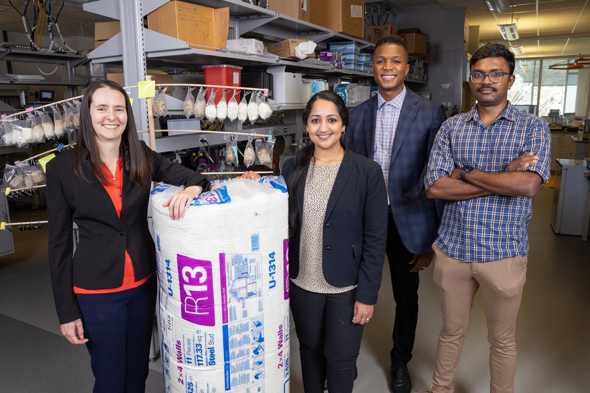 Four researchers standing in a lab with a large roll of fiberglass insulation and a wooden rack holding small bags of hemp fiber-based insulation materials. (Photo: Candler Hobbs)