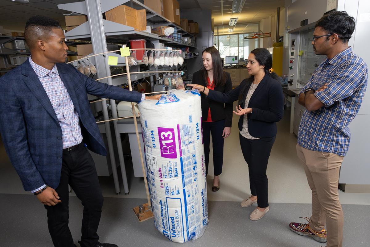 Four researchers talking in a lab. They're standing with a large roll of fiberglass insulation and a wooden rack holding small bags of prototype hemp fiber-based insulation materials. (Photo: Candler Hobbs)