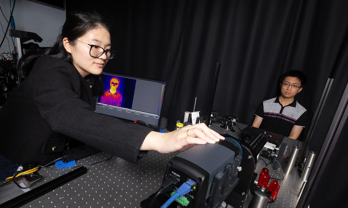 Dingding Han adjusts an infrared camera pointed at a test subject sitting in front of a black curtain. On a computer screen to her left is a thermal image of the subject. (Photo: Candler Hobbs)