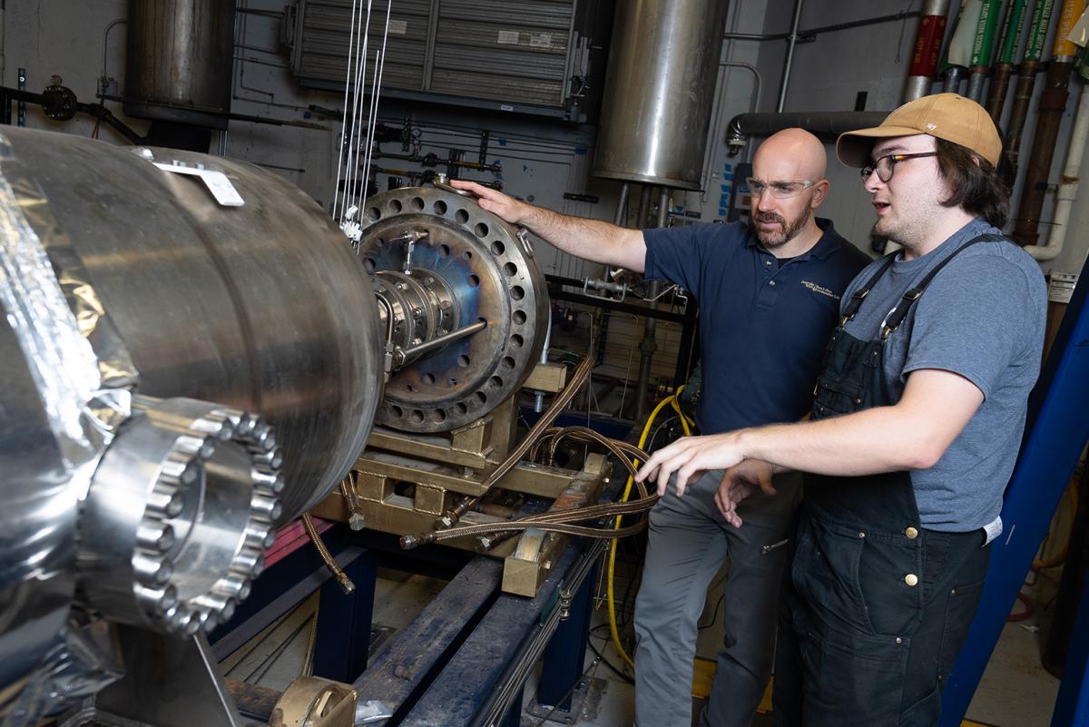 Two people standing next to a long cylindrical combustion chamber in a lab