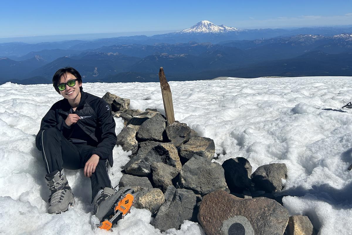 James Shin sits next to a pile of rocks in the snow atop Mt. Adams in the state of Washington with a mountain peak in the far distance behind him.