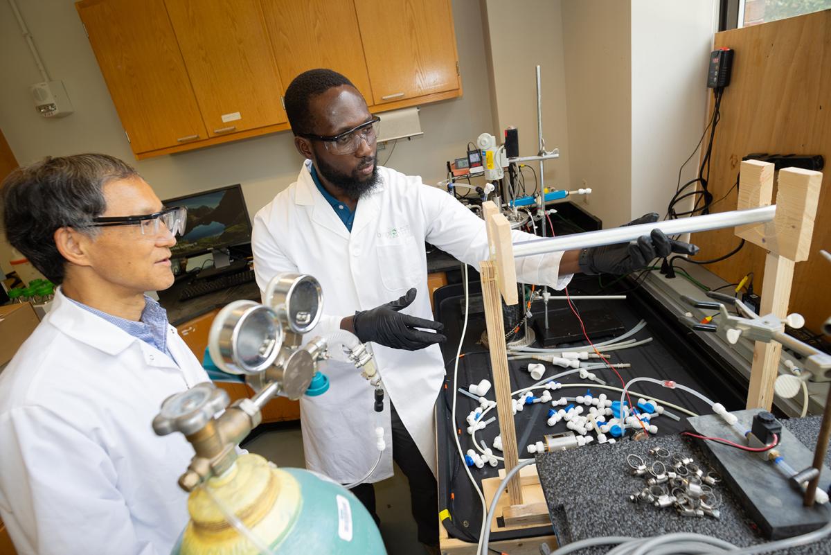 Two people discuss an experiment in a lab while looking at a table with piping and a wooden stand holding a horizontal pipe.