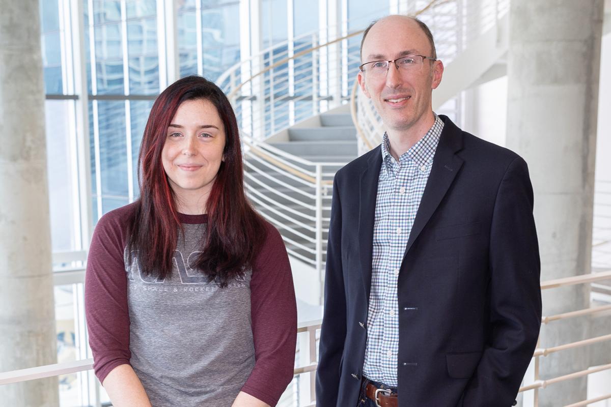 Two people stand in an atrium with stairs behind them