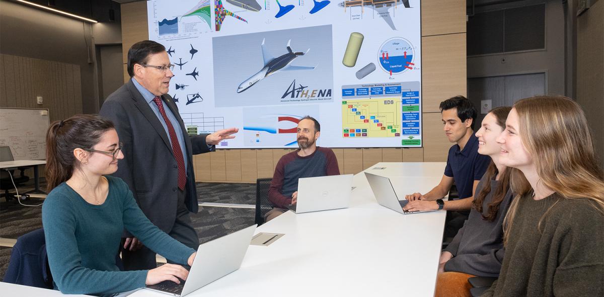 a standing person speaks to five people at a table with airplane schematics on the wall behind them