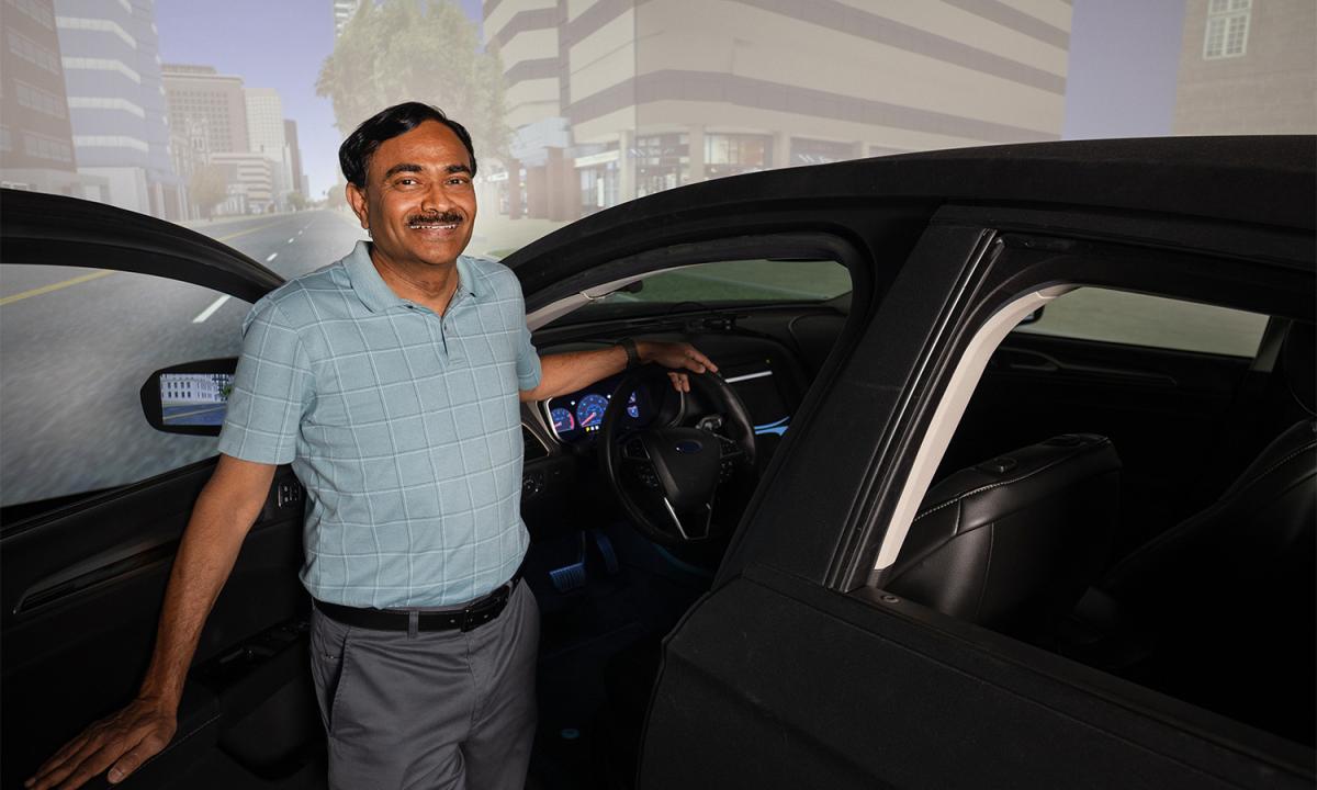 a man stands next to a car in a lab with a virtual environment on screen behind him