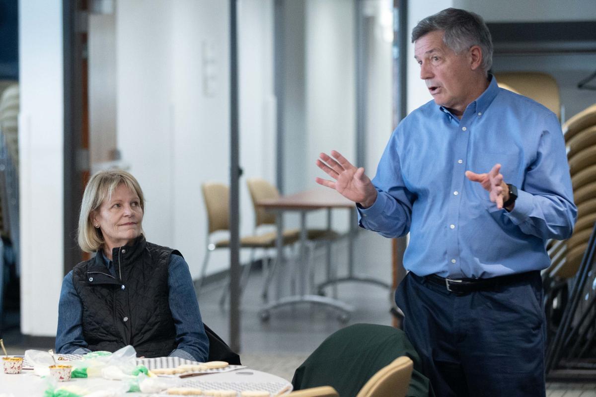 one man standing while a woman sits next to him at a table