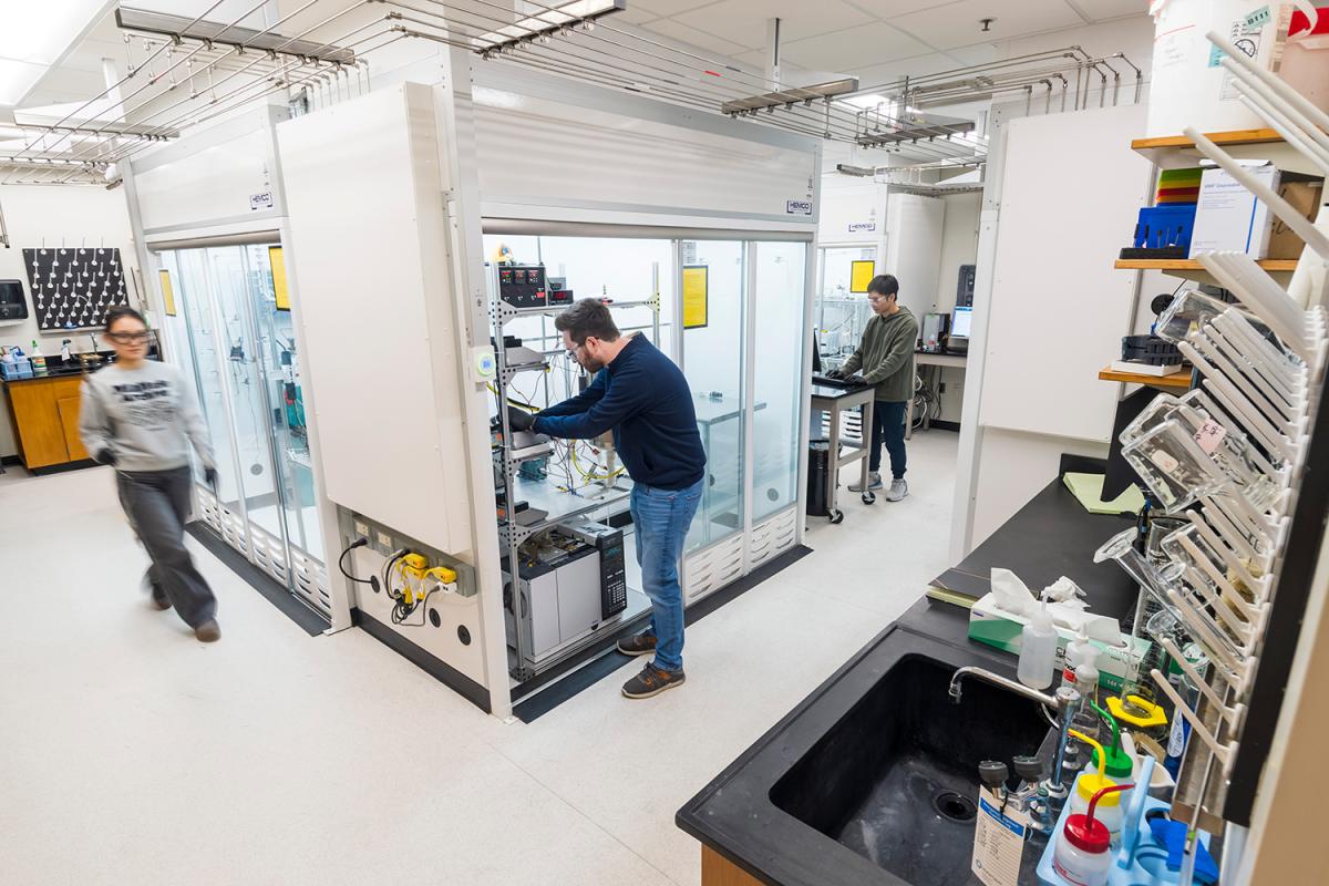 a corner view of three people working in a lab with catalyst reactor chambers and lab glassware in the foreground