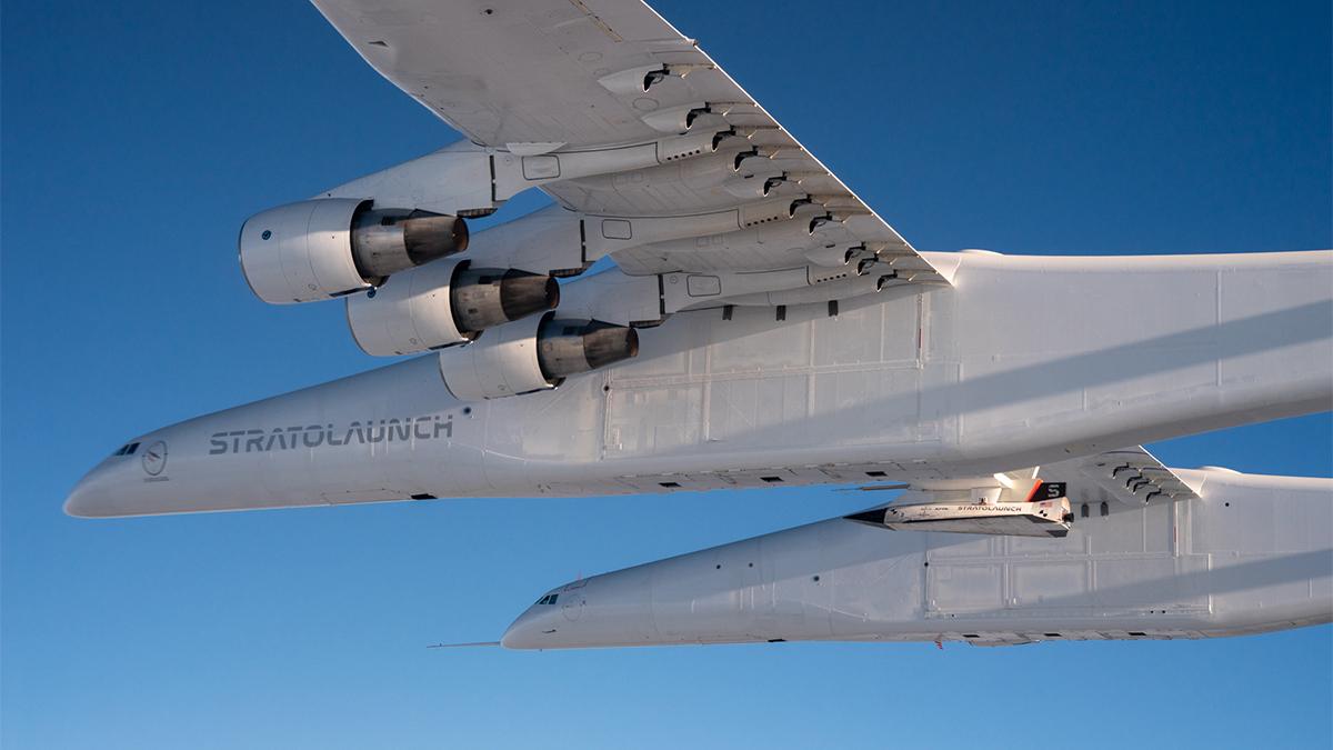 A large airplane carrying a hypersonic aircraft beneath it against a blue sky