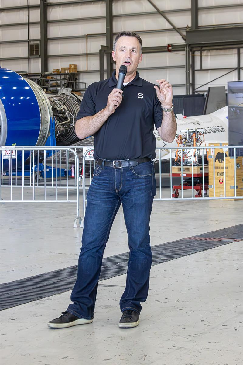 Zachary Krevor speaks with a microphone in a hangar with aircraft behind him