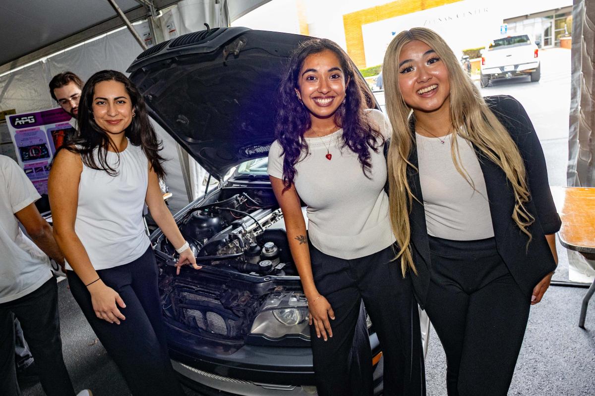 three women stand in front of a car with its hood in the air