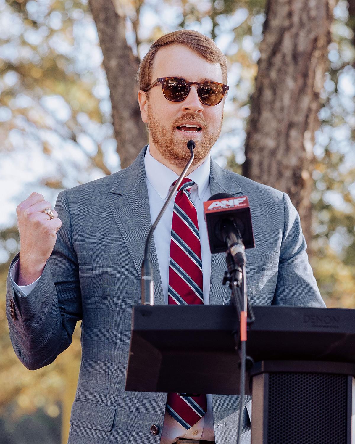 A man in a suit speaks at a podium with media microphones