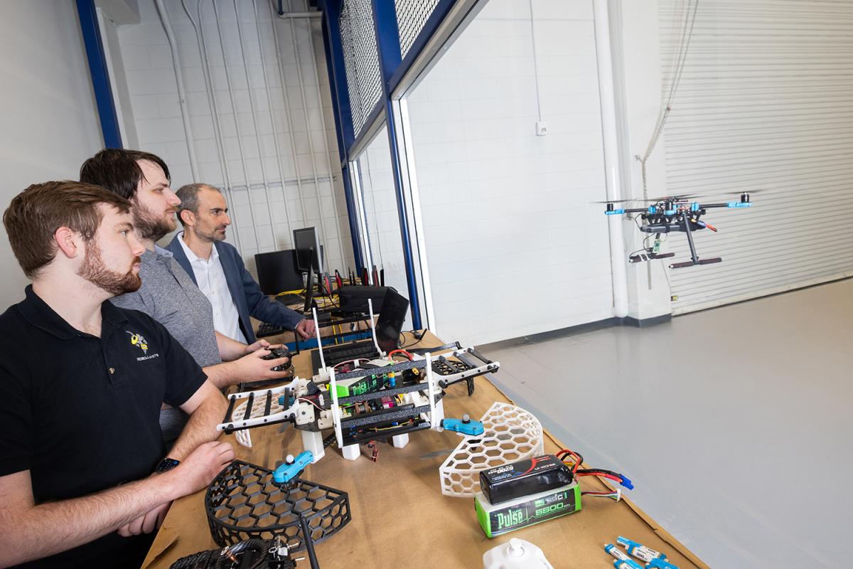 three people fly a drone on the other side of a window with more drones on a table