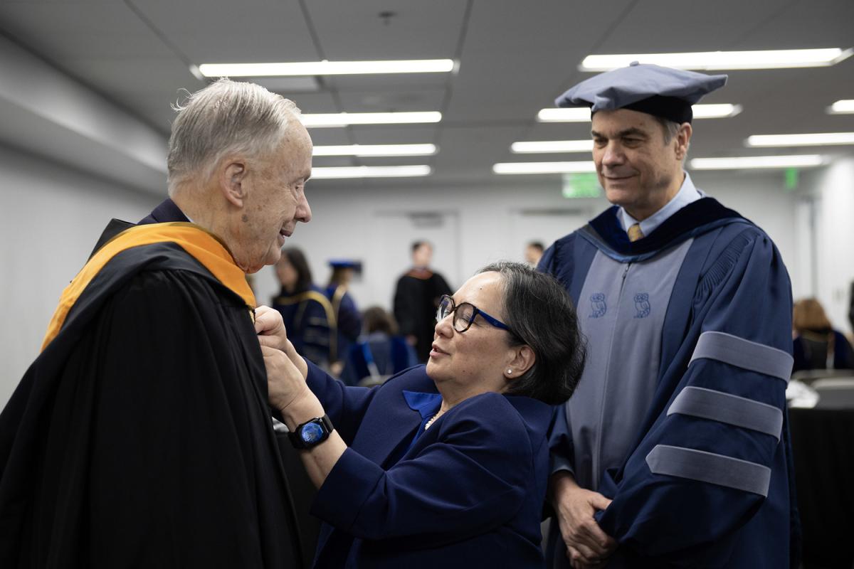 Mila Lynne Floro helps Kenneth Grundborg put on his graduation gown and master's hood.