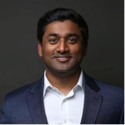 Shan Pesaru in jacket and white shirt, looking at camera with dark background